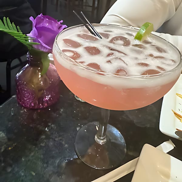 Pink cocktail with ice and a lime wedge, next to a purple rose in a vase.