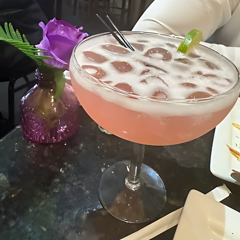 Pink cocktail with ice and a lime wedge, next to a purple rose in a vase.