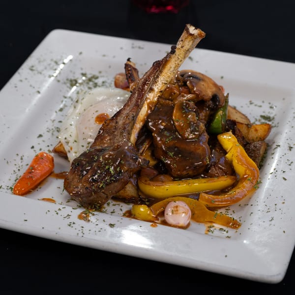 Lamb chops with fried egg, bell peppers, mushrooms, and carrots on a white plate.