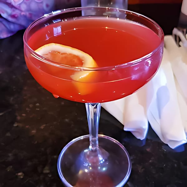 A vibrant red cocktail with a lemon slice in a coupe glass on a dark table.